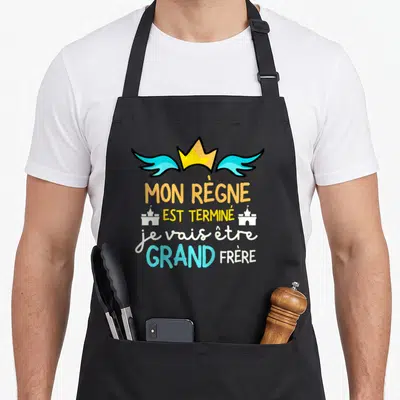 Tablier de cuisine noir avec une annonce de naissance de grand frère en lettres colorées et une couronne.