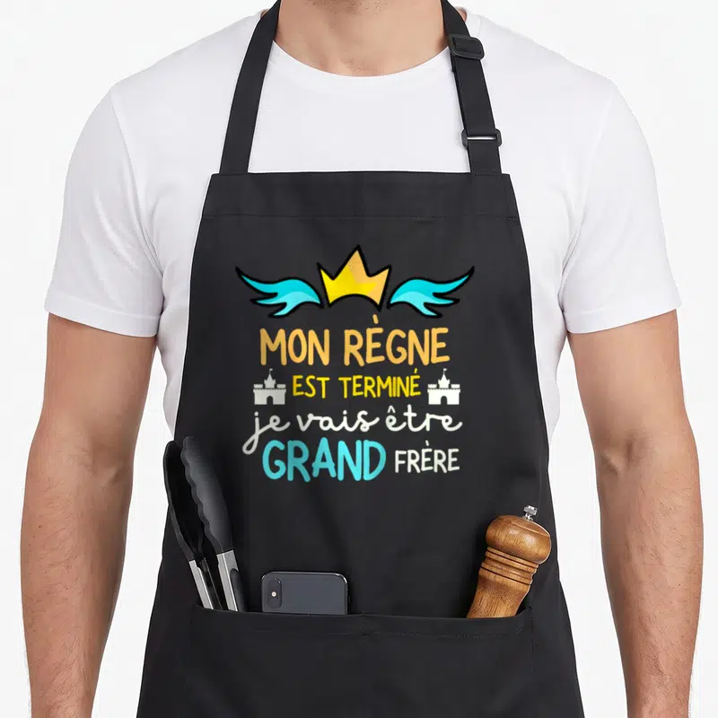 Tablier de cuisine noir avec une annonce de naissance de grand frère en lettres colorées et une couronne.