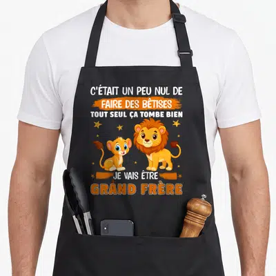 Tablier de cuisine noir avec Simba, Scar et le texte Je vais être grand frère imprimé en orange et blanc.