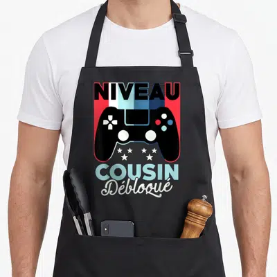 Tablier de cuisine noir avec manette de jeu et texte Niveau Cousin Débloqué imprimé en couleurs vives.