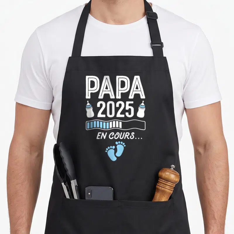 Tablier de cuisine noir avec l'inscription Papa 2025 en cours, deux biberons et des petites pattes bleues.