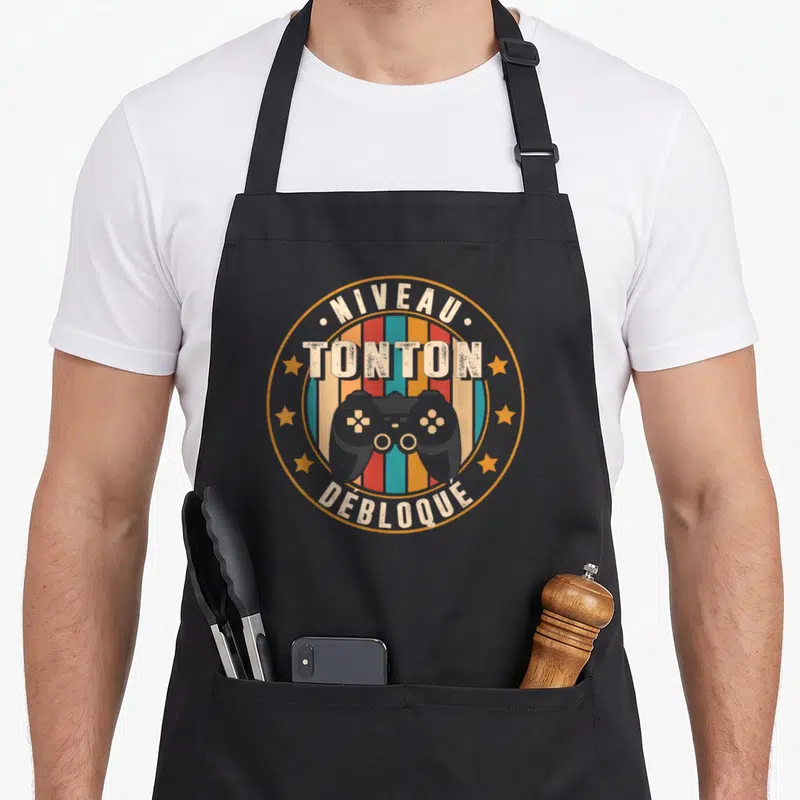 Tablier de cuisine noir avec logo circulaire Niveau Tonton Débloqué et manette de jeu vidéo grise sur fond rayé coloré.