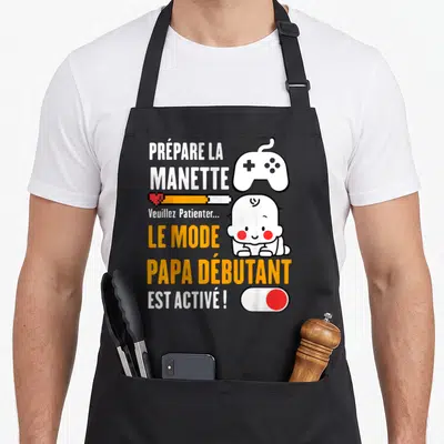 Tablier de cuisine noir avec illustration de bébé gamer et texte blanc orange sur fond uni.