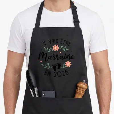 Tablier de cuisine noir avec inscription Je vais être Marraine en 2026 entouré de fleurs et d'empreintes de pieds.
