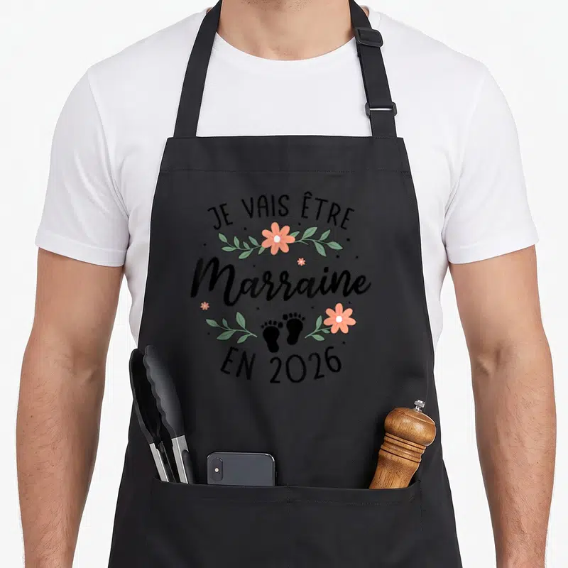 Tablier de cuisine noir avec inscription Je vais être Marraine en 2026 entouré de fleurs et d'empreintes de pieds.