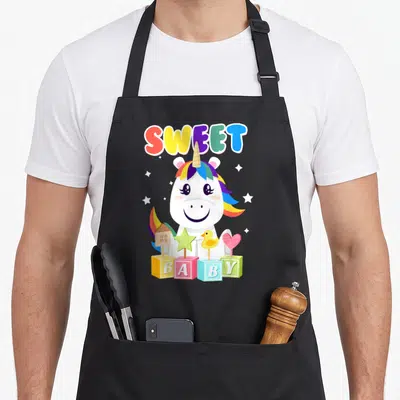 Tablier de cuisine noir avec une licorne blanche souriante, des blocs colorés formant le mot Baby et l'inscription Sweet en lettres multicolores.
