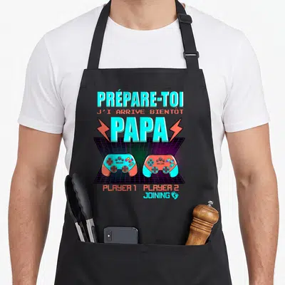 Tablier de cuisine noir avec l'inscription Prépare-toi Papa et deux manettes de jeu vidéo colorées sur fond rétro.