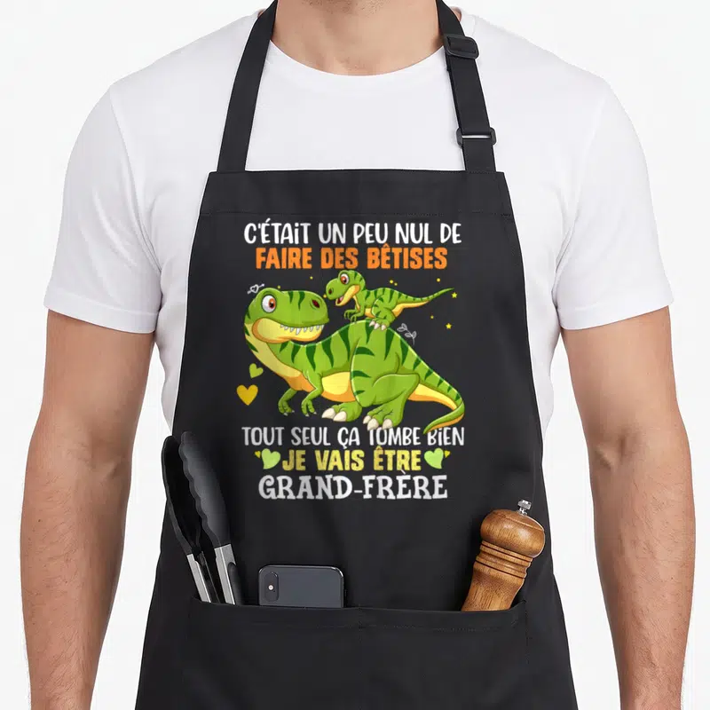 Tablier de cuisine noir avec dinosaures verts et texte annonçant la naissance d'un grand-frère.