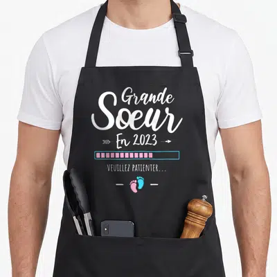 Tablier de cuisine noir avec l'inscription Grande Sœur En 2023, barre de chargement rose et deux petits pieds colorés en bas du motif.