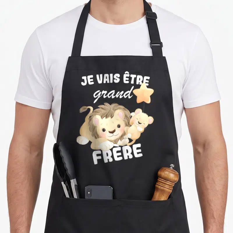 Tablier de cuisine noir avec inscription Je vais être grand frère 2026 et deux lions en dessin animé tenant une étoile.