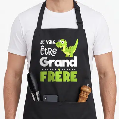 Tablier de cuisine noir avec le texte Je vais être grand frère et un petit dinosaure vert mignon imprimé au centre.