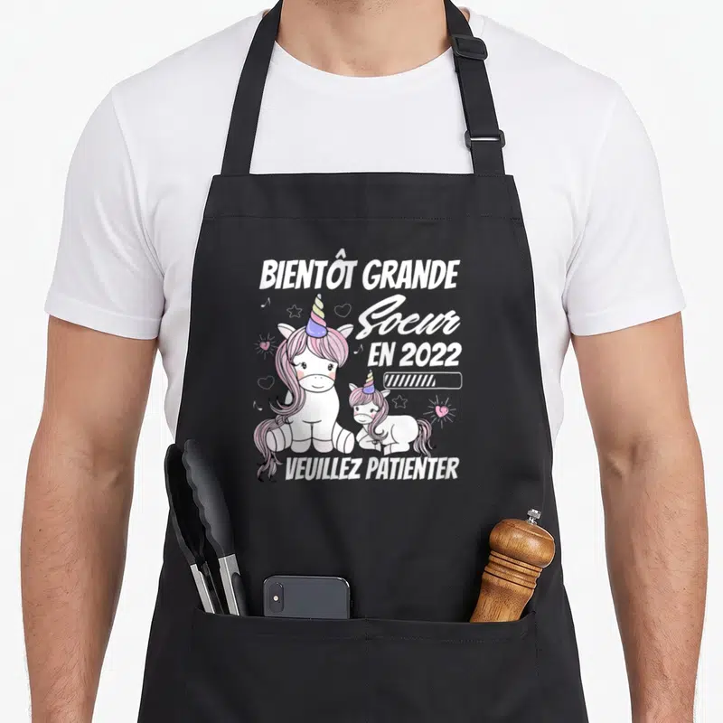 Tablier de cuisine noir avec deux licornes roses et le texte Bientôt Grande Soeur en 2022 veuillez patienter