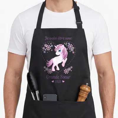 Tablier de cuisine noir avec une licorne rose, des fleurs et le texte Je vais être une grande soeur 2021.