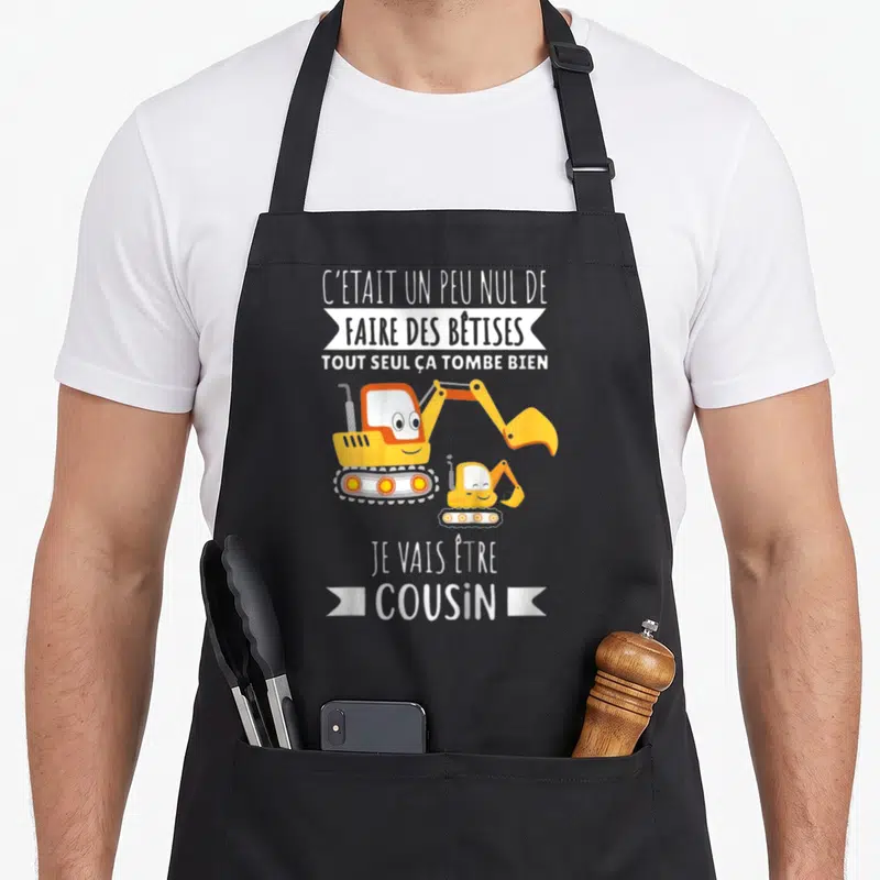 Tablier de cuisine noir avec deux pelles jaunes souriantes et le texte Je vais être cousin imprimé en blanc.