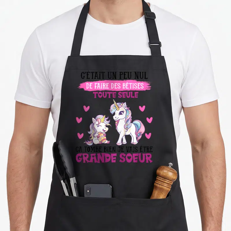 Tablier de cuisine noir avec deux licornes roses et texte annonçant le rôle de grande sœur.