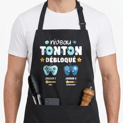 Tablier de cuisine noir avec deux manettes de jeu vidéo bleues et l'inscription Niveau Tonton Débloqué en lettres blanches et bleues.