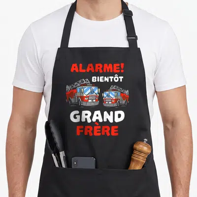 Tablier de cuisine noir avec deux camions de pompiers rouges et le texte Alarme Bientôt Grand Frère imprimé en rouge et blanc.