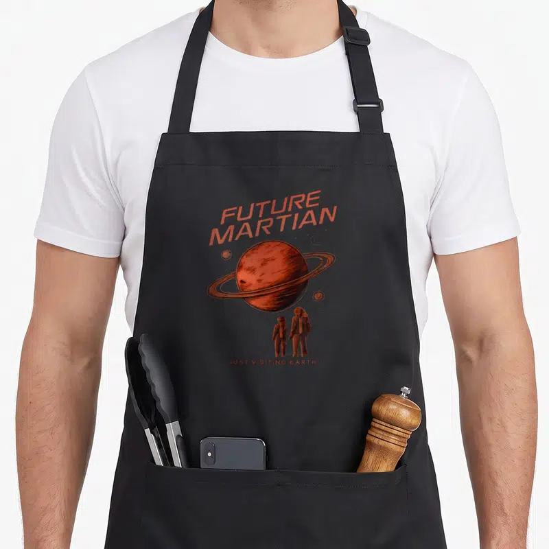Tablier de cuisine noir avec illustration d'une planète rouge et deux enfants sous le texte Future Martian Just Visiting Earth en orange.