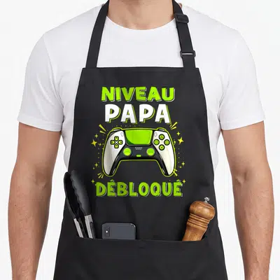 Tablier de cuisine noir avec une manette de jeu vidéo verte et blanche et le texte Niveau Papa Débloqué.