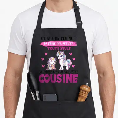 Tablier de cuisine noir avec deux licornes roses et le texte Je vais être cousine en lettres violettes et roses.
