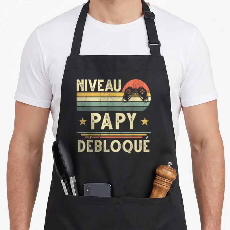 Tablier de cuisine noir avec inscription Niveau Papy Débloqué et illustration de manette de jeu vidéo style rétro coloré.