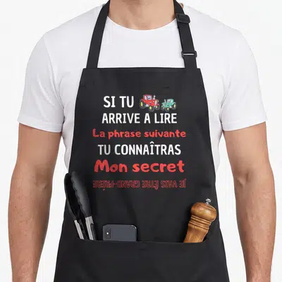 Tablier de cuisine noir avec texte blanc et rouge annonçant un secret de grossesse au-dessus de deux tracteurs colorés.