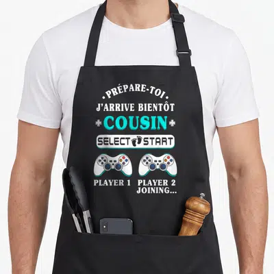 Tablier de cuisine noir avec texte blanc et turquoise Prépare-toi j'arrive bientôt cousin Select Start Player 1 Player 2 joining et deux manettes de jeu vidéo.