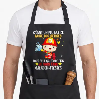 Tablier de cuisine noir avec un pompier cartoon et le message Je vais être grand-frère imprimé en couleurs vives.