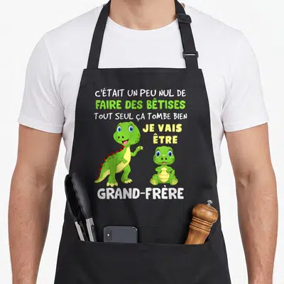 Tablier de cuisine noir avec deux dinosaures verts et le texte Je vais être grand-frère imprimé en centre.