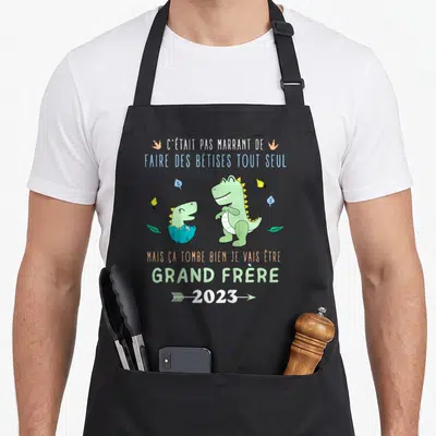 Tablier de cuisine noir avec deux dinosaures verts et le texte annonçant un futur grand frère en 2023.