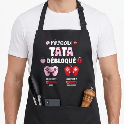 Tablier de cuisine noir avec les mots Niveau Tata Débloqué, deux manettes de jeu roses et rouges et des cœurs stylisés.