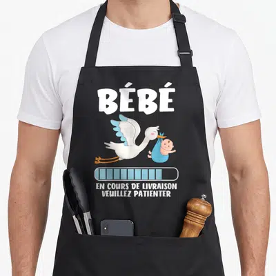 Tablier de cuisine noir avec illustration cigogne portant bébé et texte en cours de « livraison » veuillez patienter.