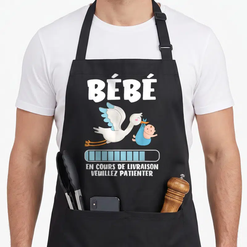 Tablier de cuisine noir avec illustration cigogne portant bébé et texte en cours de « livraison » veuillez patienter.