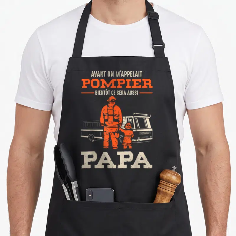 Tablier de cuisine noir avec illustration orange d'un pompier et un enfant devant un camion, texte blanc et orange.