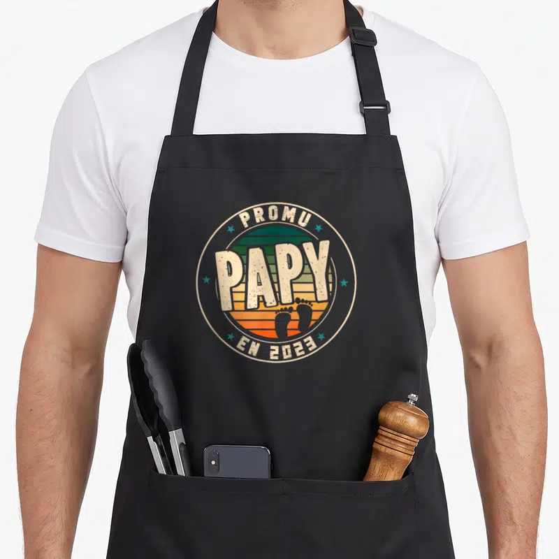 Tablier de cuisine noir avec un logo circulaire Promu Papy en 2023 sur fond de soleil couchant et empreintes de pieds.