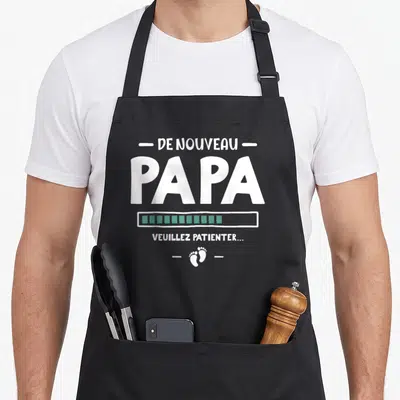 Tablier de cuisine noir avec texte blanc De nouveau Papa, barre de progression verte et empreintes de pieds.