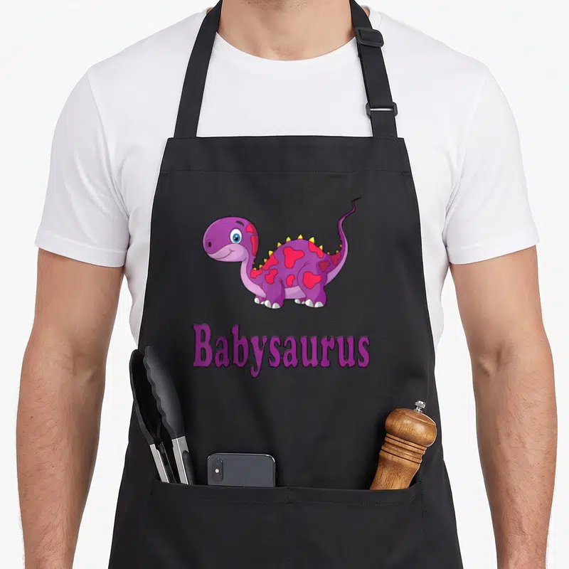 Tablier de cuisine noir avec un dinosaure violet mignon portant des cœurs roses et l'inscription Babysaurus.
