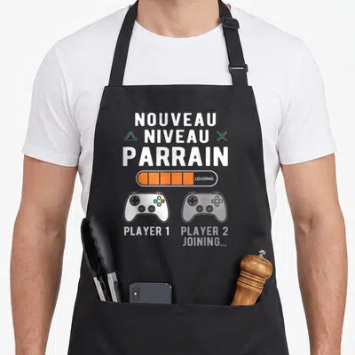 Tablier de cuisine noir avec l'inscription Nouveau Niveau Parrain et deux manettes de jeu vidéo Player 1 et Player 2 joining.