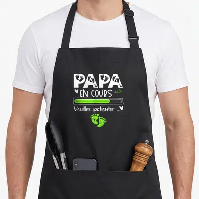 Tablier de cuisine noir avec l'inscription Papa en cours et des empreintes de pieds vertes stylisées.