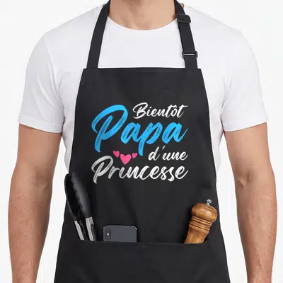 Tablier de cuisine noir avec le texte Bientôt Papa d'une Princesse en bleu et blanc orné de trois cœurs roses