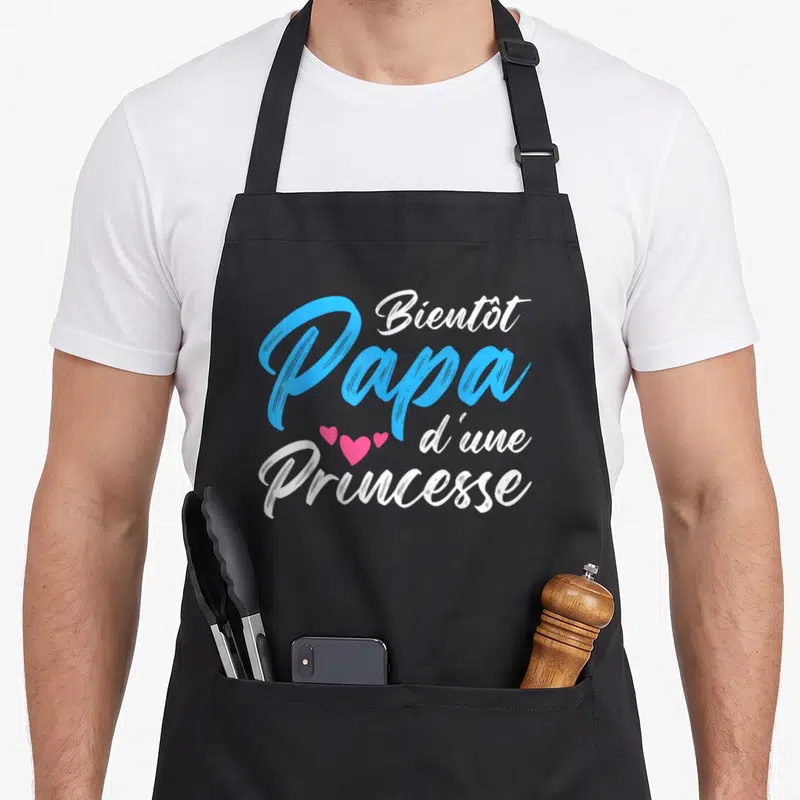 Tablier de cuisine noir avec le texte Bientôt Papa d'une Princesse en bleu et blanc orné de trois cœurs roses