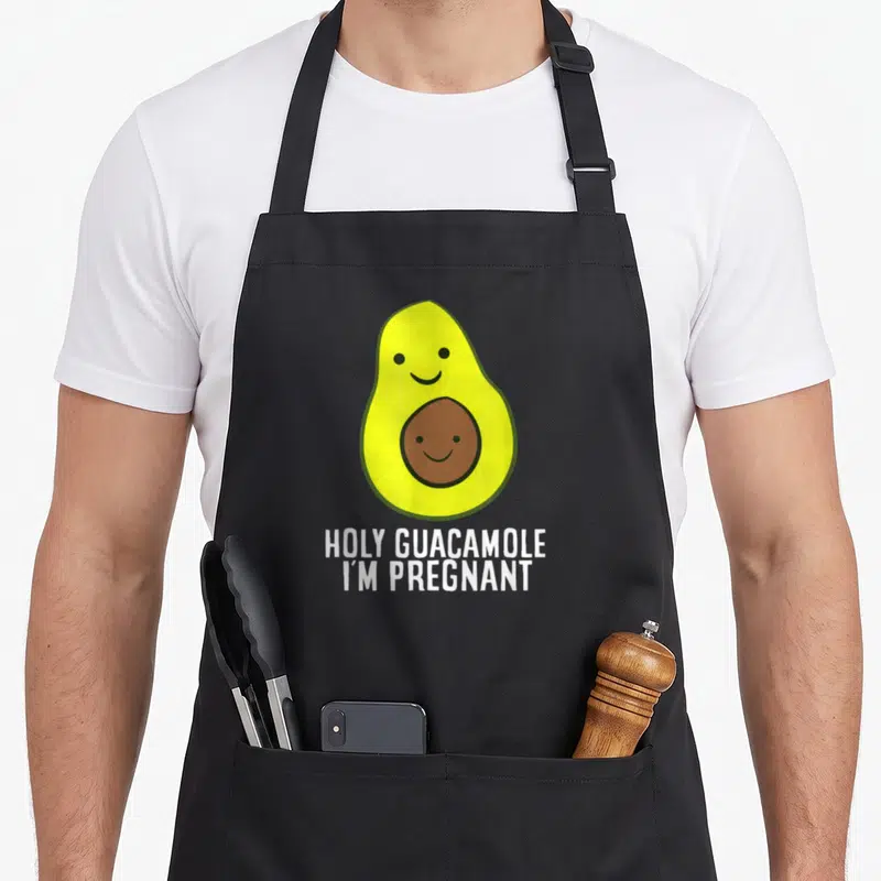 Tablier de cuisine noir avec un avocat souriant et une noyau à visage, sous le texte Holy Guacamole I'm Pregnant en blanc.
