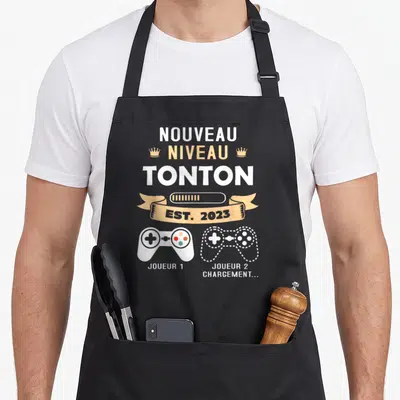 Tablier de cuisine noir avec texte Nouveau Niveau Tonton Est 2023 et deux manettes de jeu vidéo Joueur 1 et Jouleur 2 Chargement.