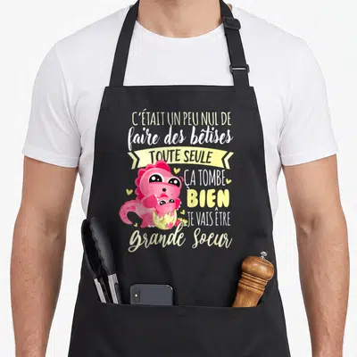 Tablier de cuisine noir avec dinosaure rose tenant un bébé et texte humoristique sur la future grande sœur.