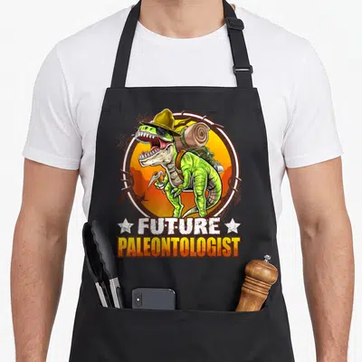 Tablier de cuisine noir avec illustration d'un dinosaure vert en explorateur portant un chapeau jaune et tenant un outil de paléontologie.