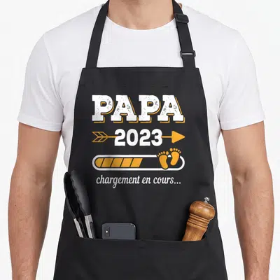 Tablier de cuisine noir avec inscription Papa 2023 chargement en cours et empreintes de pieds oranges.
