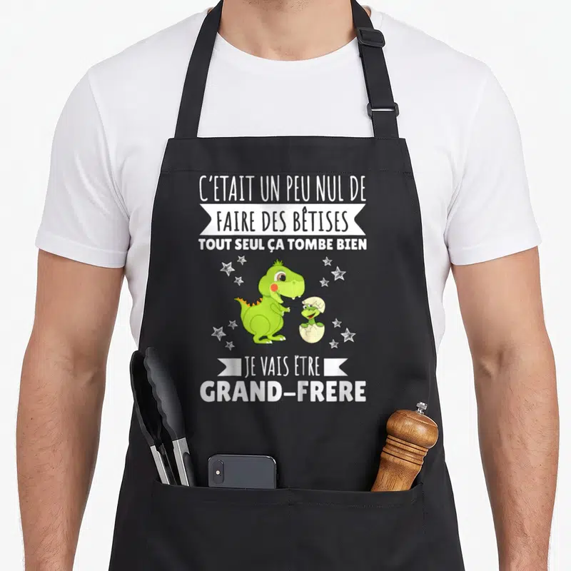 Tablier de cuisine noir avec dinosaure vert tenant un bébé dinosaure dans une coquille, entouré d'étoiles et texte blanc humoristique.