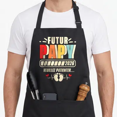 Tablier de cuisine noir avec inscription Futur Papy 2026 et petits pieds imprimés en couleur.