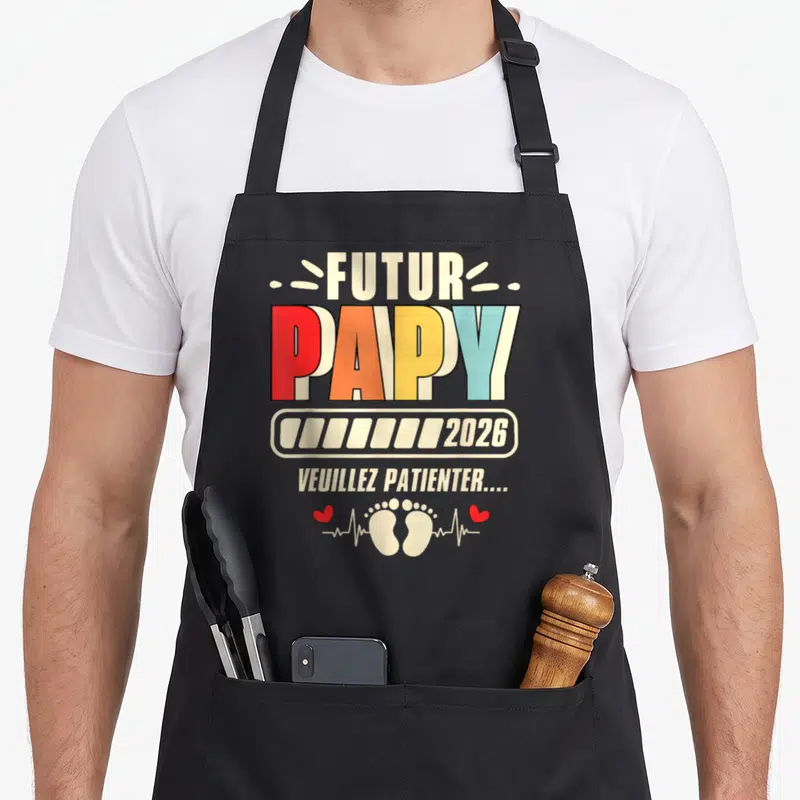 Tablier de cuisine noir avec inscription Futur Papy 2026 et petits pieds imprimés en couleur.