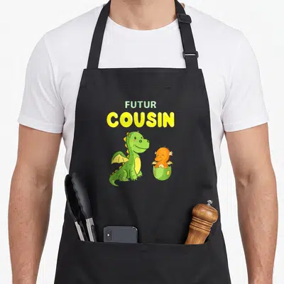 Tablier de cuisine noir avec deux dinosaures colorés et le texte Futur Cousin imprimé en jaune et vert clair.
